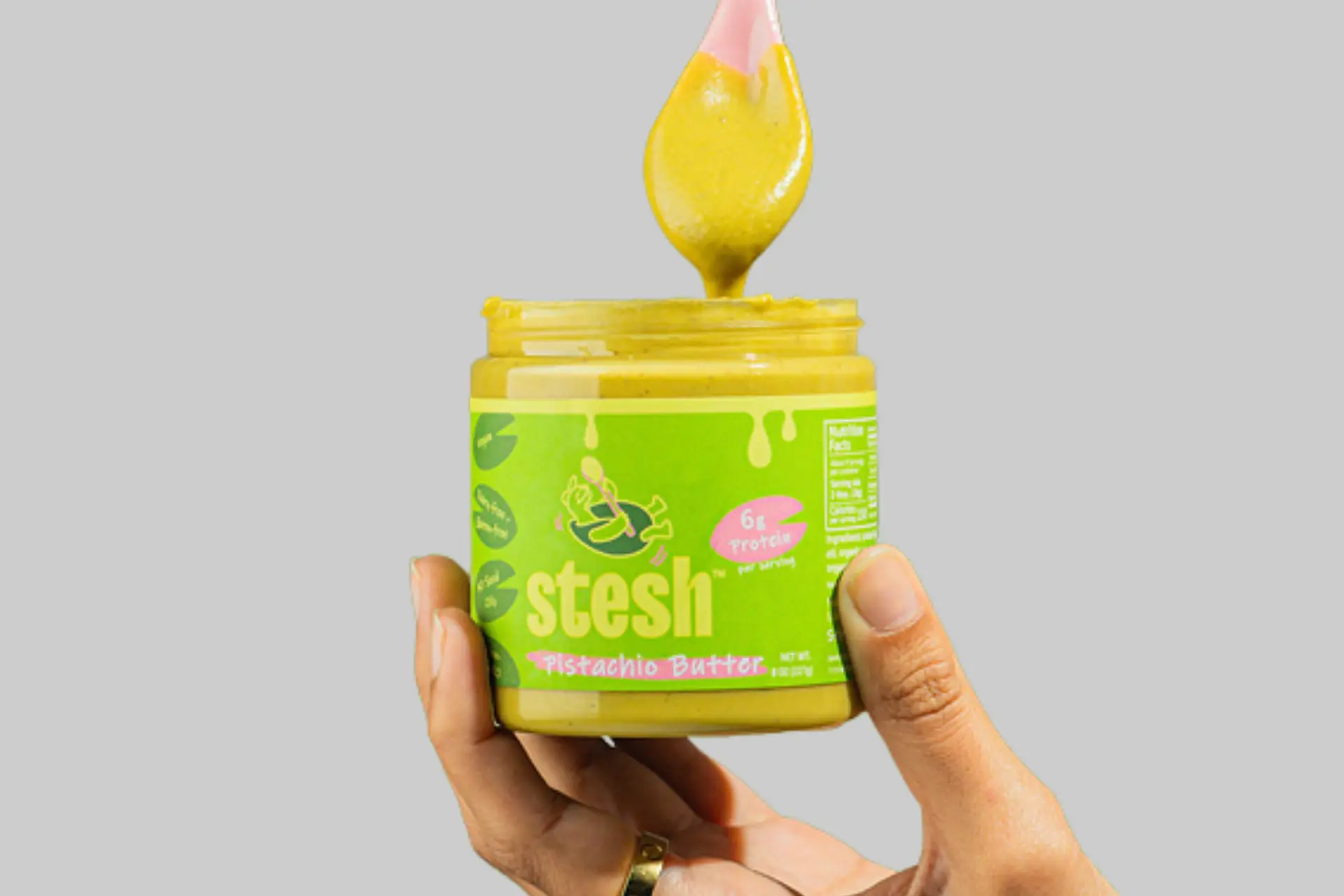 A jar of Stesh Pistachio Butter with its vibrant green color, shown with a spoon dipping in, surrounded by fresh fruit and toast. - Product Review